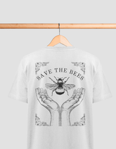 Das Bild zeigt ein weißes T-Shirt auf einem Holzbügel, das an einer Holzstange hängt. Auf dem Rücken des T-Shirts befindet sich eine Illustration mit der Aufschrift „SAVE THE BEES“. Darunter sind zwei geöffnete Hände abgebildet, die eine Biene halten. Das
