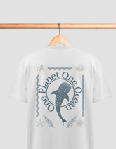 Das Bild zeigt ein weißes T-Shirt auf einem Holzbügel, das an einer Holzstange hängt. Auf dem Rücken des T-Shirts befindet sich ein blaues Design mit der Aufschrift „One Planet One Ocean“. In der Mitte ist ein Walhai abgebildet, umgeben von Wellenlinien u