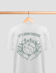 Das Bild zeigt ein weißes T-Shirt, das an einem Holzbügel hängt. Auf der Rückseite des T-Shirts ist ein weißer Aufdruck mit dem Text "SAVE THE BEES". Im Mittelpunkt des Designs befindet sich eine detaillierte Darstellung einer Biene, die über zwei geöffnete, nach oben gerichtete Hände schwebt. In den Ecken des Designs sind florale Muster abgebildet, die die Natur und den Umweltschutz symbolisieren.