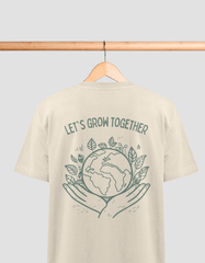 Das Bild zeigt ein beiges T-Shirt, das an einem Holzbügel hängt. Auf der Rückseite des T-Shirts ist ein weißer Aufdruck mit dem Text "SAVE THE BEES". Im Mittelpunkt des Designs befindet sich eine detaillierte Darstellung einer Biene, die über zwei geöffnete, nach oben gerichtete Hände schwebt. In den Ecken des Designs sind florale Muster abgebildet, die die Natur und den Umweltschutz symbolisieren.