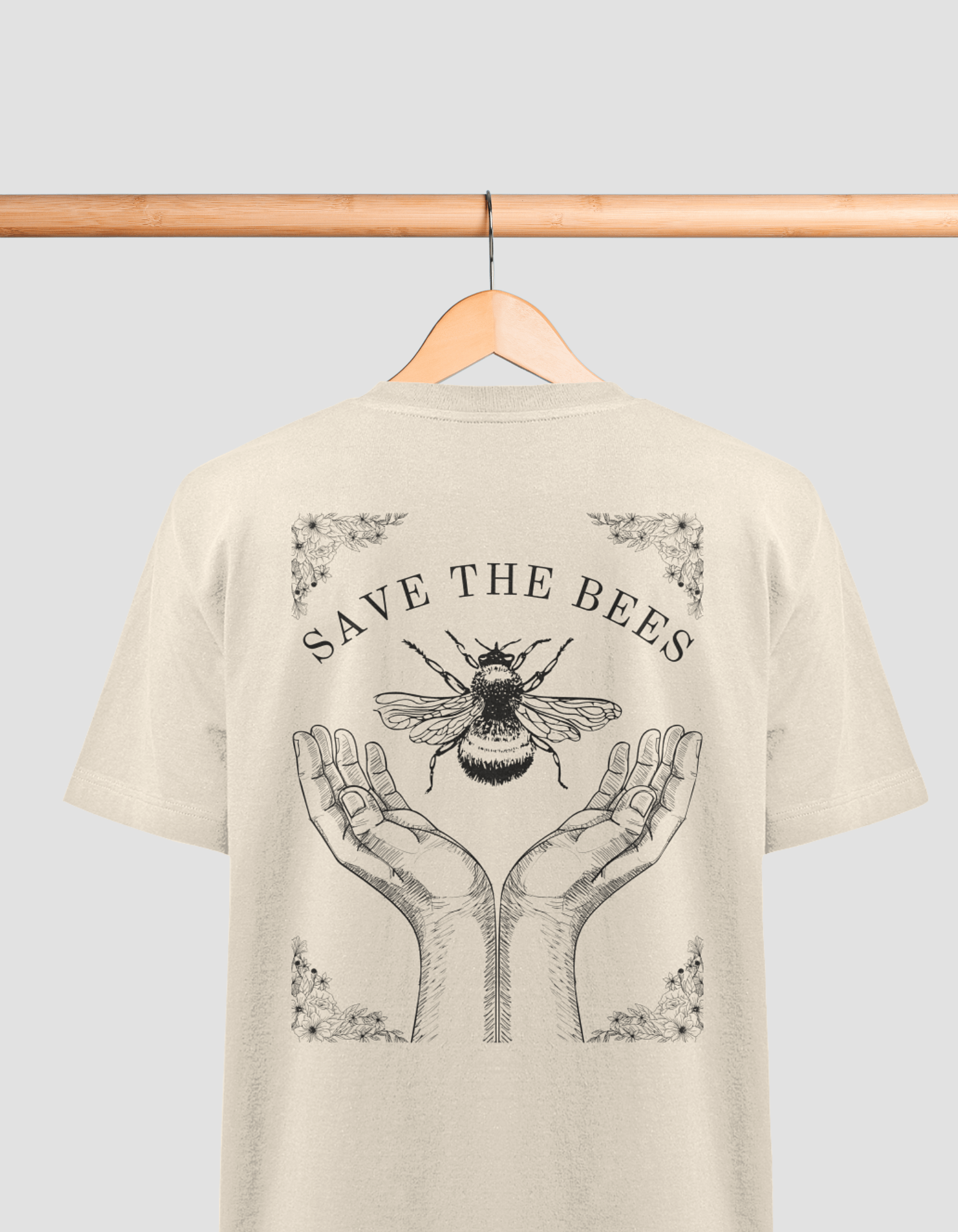 Das Bild zeigt ein beiges T-Shirt auf einem Holzbügel, das an einer Holzstange hängt. Auf dem Rücken des T-Shirts befindet sich eine Illustration mit der Aufschrift „SAVE THE BEES“. Darunter sind zwei geöffnete Hände abgebildet, die eine Biene halten. Das
