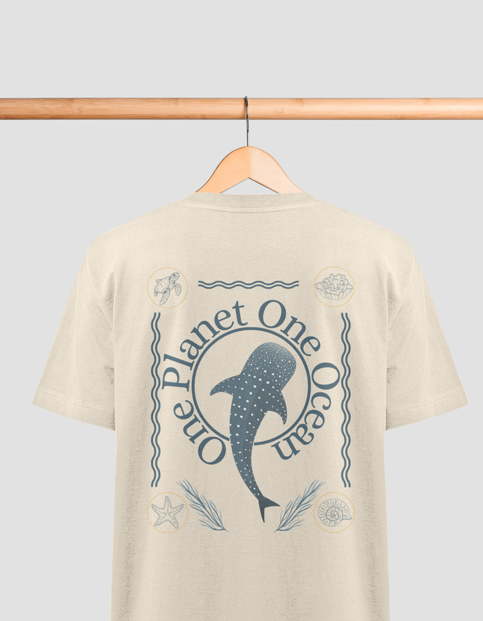 Das Bild zeigt ein beiges T-Shirt auf einem Holzbügel, das an einer Holzstange hängt. Auf dem Rücken des T-Shirts befindet sich ein blaues Design mit der Aufschrift „One Planet One Ocean“. In der Mitte ist ein Walhai abgebildet, umgeben von Wellenlinien u