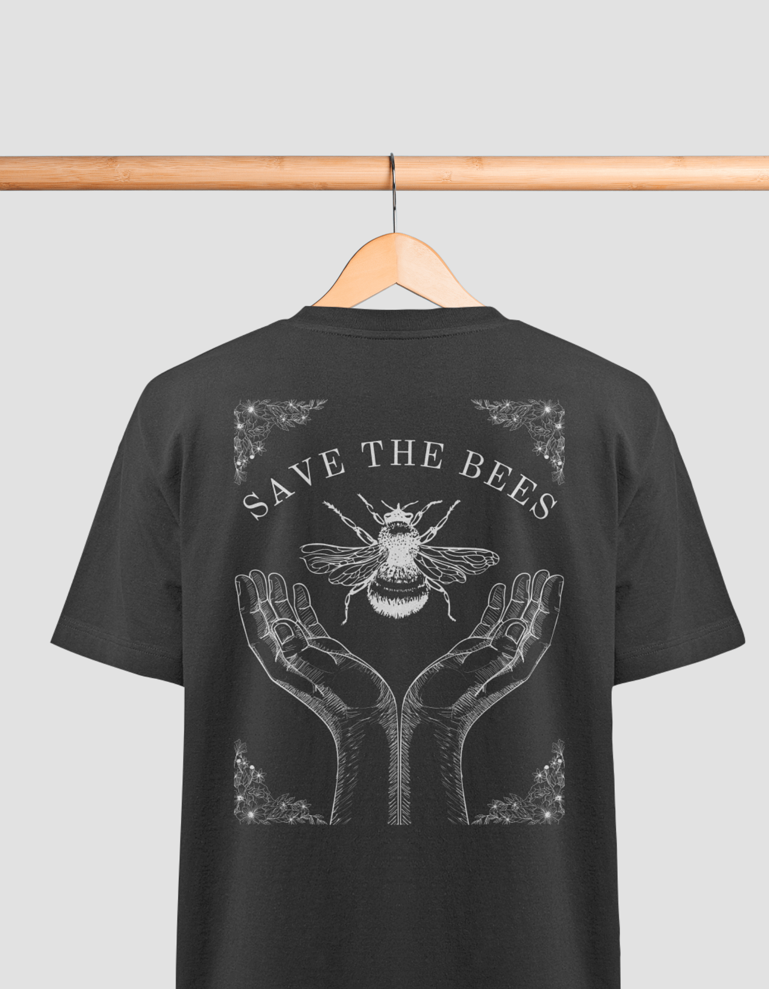 Das Bild zeigt ein schwarzes T-Shirt auf einem Holzbügel, das an einer Holzstange hängt. Auf dem Rücken des T-Shirts befindet sich eine Illustration mit der Aufschrift „SAVE THE BEES“. Darunter sind zwei geöffnete Hände abgebildet, die eine Biene halten. 